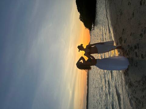       Silhouetted women in flowing skirts pose playfully on the shoreline at sunset.
  