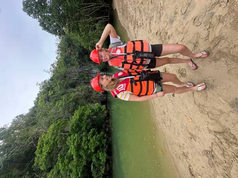       Two women wearing helmets and orange life vests stand on a sandy bank beside emerald water and lush cave entrance.
  