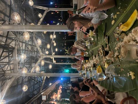       Long banana-leaf feast with travelers sharing food and drinks under decorative lights.
  