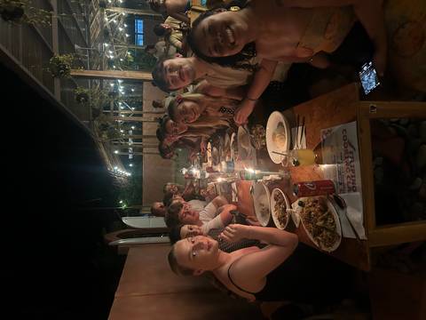       Large group of young travelers having dinner together at a long outdoor table lit by candles
  
