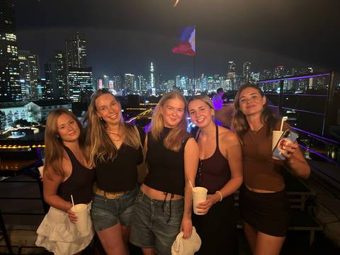       Five friends posing on a rooftop bar with a brightly lit city skyline behind them at night
  