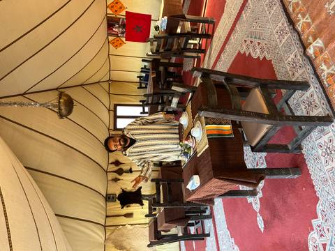       Host in striped djellaba welcomes guests inside a spacious desert camp dining tent.
  