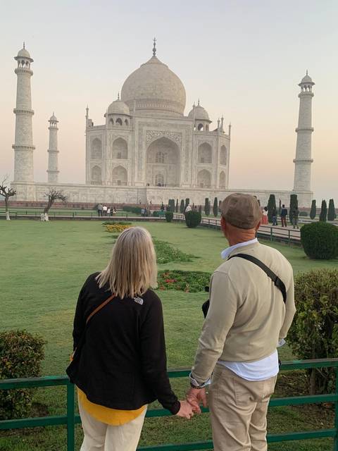       Two travellers admire the Taj Mahal from the lush morning gardens with soft sunrise light.
  
