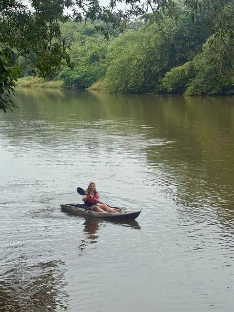       A woman paddles a kayak along a calm green river.
  