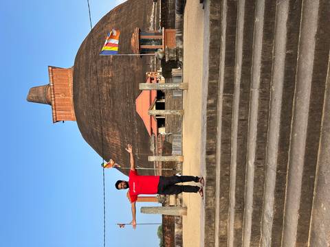       A traveler spreads his arms in front of a massive brick dagoba under blue sky.
  