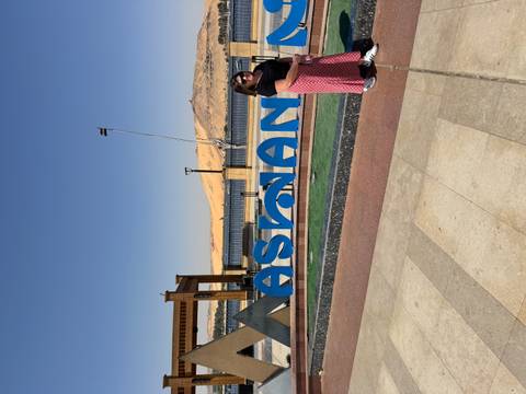       Smiling woman next to a bright blue Aswan sign with river and sand dunes in view.
  