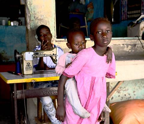       Young girl in pink dress carries a toddler while another child watches from behind a sewing machine.
  