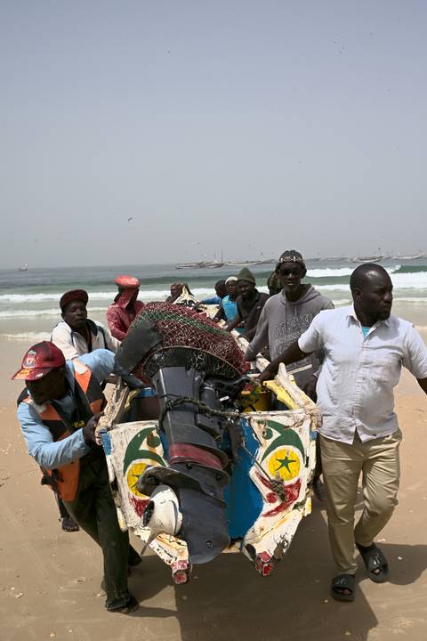       Fishermen haul a heavy engine and nets ashore on a windy beach with surf and boats offshore.
  