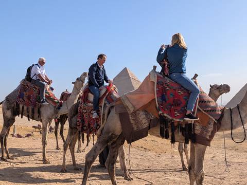       Tourists riding camels with the pyramids rising from the Giza desert in the background.
  