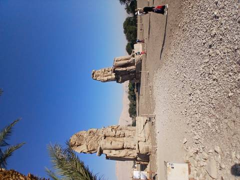       The Colossi of Memnon statues towering above visitors under a clear blue sky.
  
