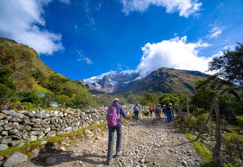       Backpackers hike a rocky trail toward snow-covered Andean peaks under a vivid blue sky.
  