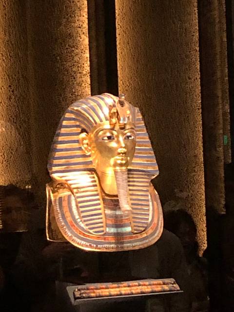       Illuminated golden funerary mask of Tutankhamun displayed in a museum setting.
  