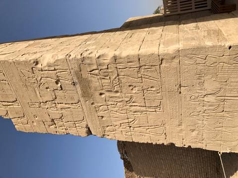       Close-up of an ancient Egyptian sandstone wall covered in detailed hieroglyphs and carved figures under a blue sky.
  