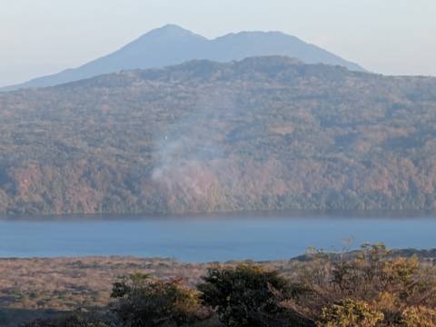       Distant smoking volcano above a blue crater lake framed by dry forest slopes.
  