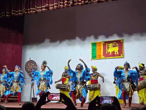       Traditional Sri Lankan dancers in blue costumes performing on stage beside the national flag.
  