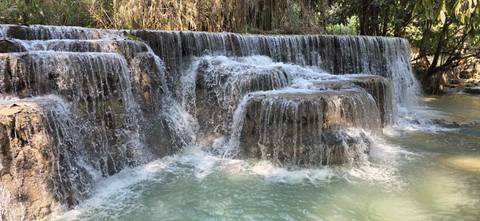       Turquoise water cascades over tiered limestone falls surrounded by lush foliage.
  