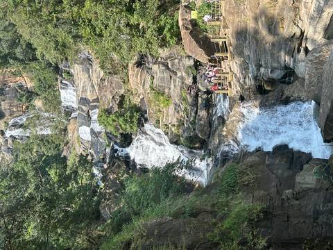       A multi-tiered waterfall tumbles through jungle cliffs as visitors gather on a viewing deck.
  
