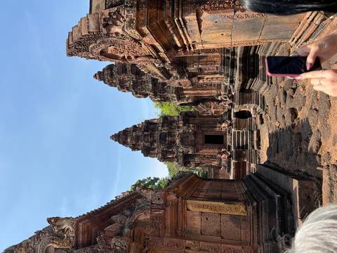      Intricately carved red sandstone towers of Banteay Srei with tourists photographing.
  