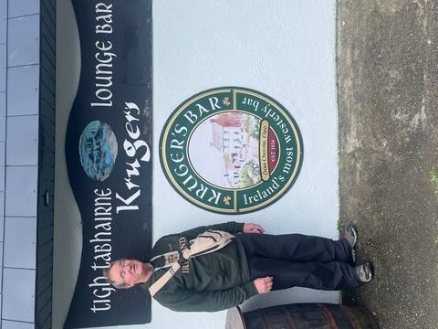       Older male traveler poses beside the exterior sign of Kruger’s Bar painted on a pale wall.
  