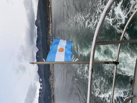       Argentinian flag flutters from a boat stern with wake, mountains and Ushuaia town under cloudy sky.
  