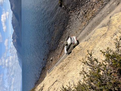       White 4x4 drives a narrow dirt track along a vast lake with snow-capped mountains in background.
  