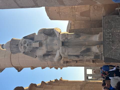       Massive seated statue of Ramses II carved in stone with tourists at its base, late afternoon light.
  