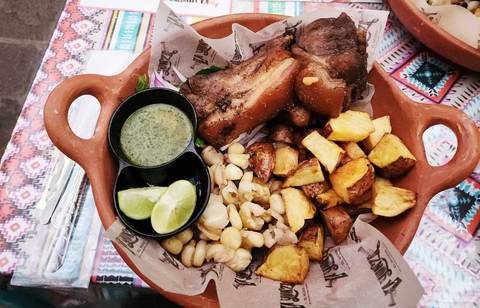       Peruvian dish featuring roasted pork, corn, potatoes, lime wedges and green sauce served in a clay bowl.
  