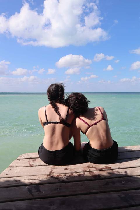      Two young women in swimwear sit side by side facing turquoise Caribbean sea under sunny skies.
  