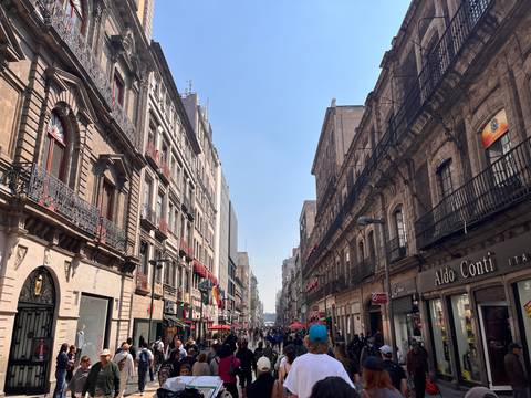       Bustling historic pedestrian street in Mexico City lined with ornate buildings, flags and shops on sunny day.
  