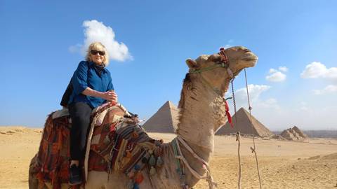       Traveler riding a camel in front of the iconic Giza pyramids beneath a vivid blue sky.
  