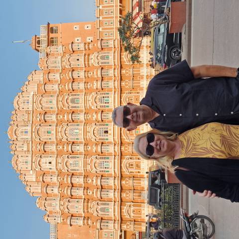       Smiling couple wearing sunglasses posing in front of the ornate pink-sandstone façade of the Hawa Mahal palace.
  