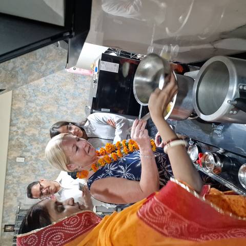       Tourist wearing a marigold garland stirs a pot during an Indian cooking lesson inside a home kitchen while others watch.
  