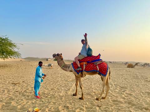       Traveler seated on a decorated camel giving a victory sign while a local handler stands beside on golden desert dunes.
  