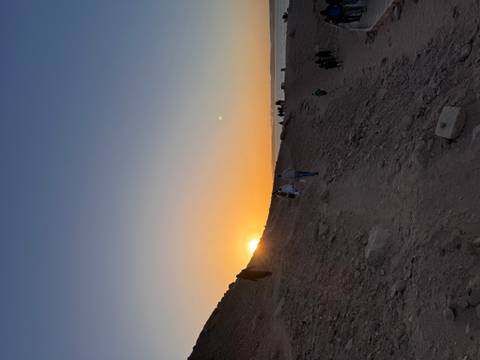       Travelers ascend a rocky path as the sun sets behind desert hills, casting an orange glow.
  