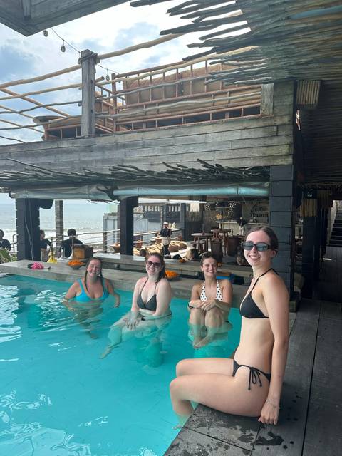       Four young women smile while relaxing in a beachfront infinity pool at a rustic bar.
  