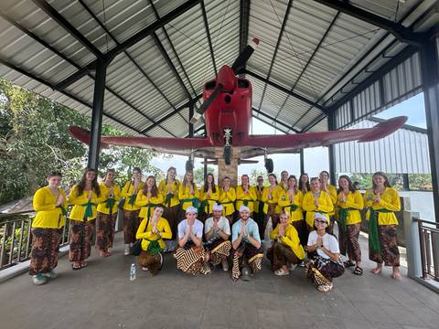       Large tour group dressed in matching yellow shirts poses beneath a red seaplane in an open-air hangar.
  