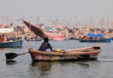       Man rows a weathered wooden boat on a wide river with a busy, colorful festival shoreline in the background.
  