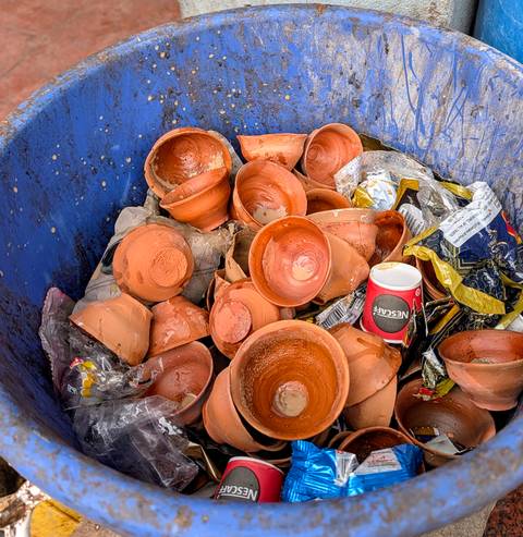       Discarded clay tea cups and assorted trash fill a blue bin, showing post-ceremony waste.
  