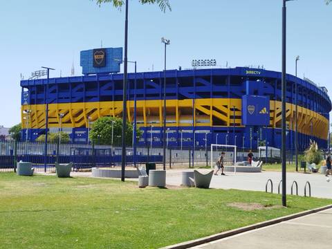       Blue and yellow La Bombonera football stadium basking in bright sunshine, viewed from a grassy park.
  