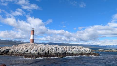       Red-and-white Les Éclaireurs lighthouse stands on a rugged rocky islet under a vivid blue sky with scattered clouds.
  