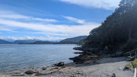       Calm blue channel edged by rocky shoreline with distant snow-capped peaks beneath a partly cloudy sky.
  