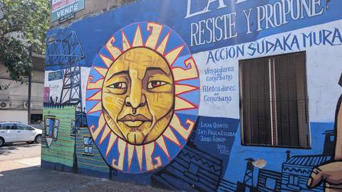       Vibrant street mural featuring a stylized sun face and Spanish slogans on a blue wall in La Boca.
  