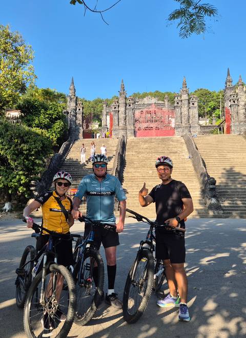       Three cyclists pose with bikes before the grand stone staircase of Khai Dinh Tomb.
  