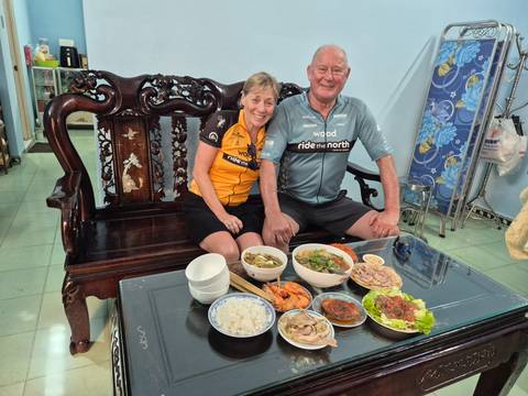       Couple relax indoors behind a spread of Vietnamese home cooking laid out on a glass table.
  