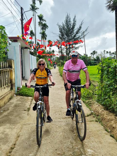       Two cyclists ride a rural concrete lane with lush green rice paddies on either side.
  