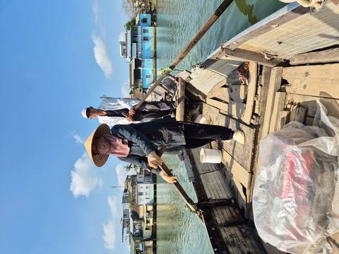      Traditional wooden boat propelled by a standing rower carries a passenger across a calm channel.
  