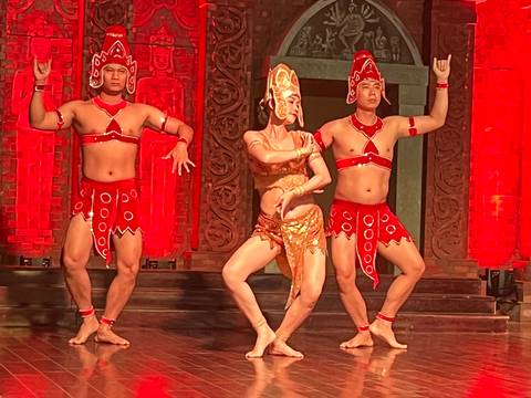       Three dancers in red and gold costumes perform an energetic traditional routine on a stage lit with red lighting.
  