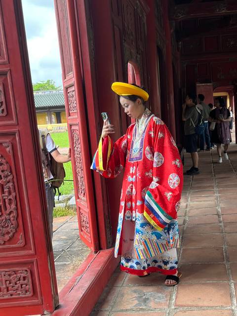       Woman in ornate royal costume takes a selfie inside a richly carved red wooden corridor.
  