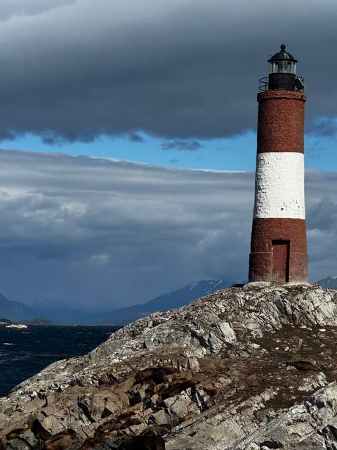       Red-and-white striped lighthouse standing on a rocky outcrop with dramatic clouds and snowy mountains in the background.
  