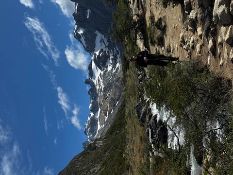       Hiker on a rocky trail beside a rushing stream with jagged snowy peaks and blue sky overhead.
  